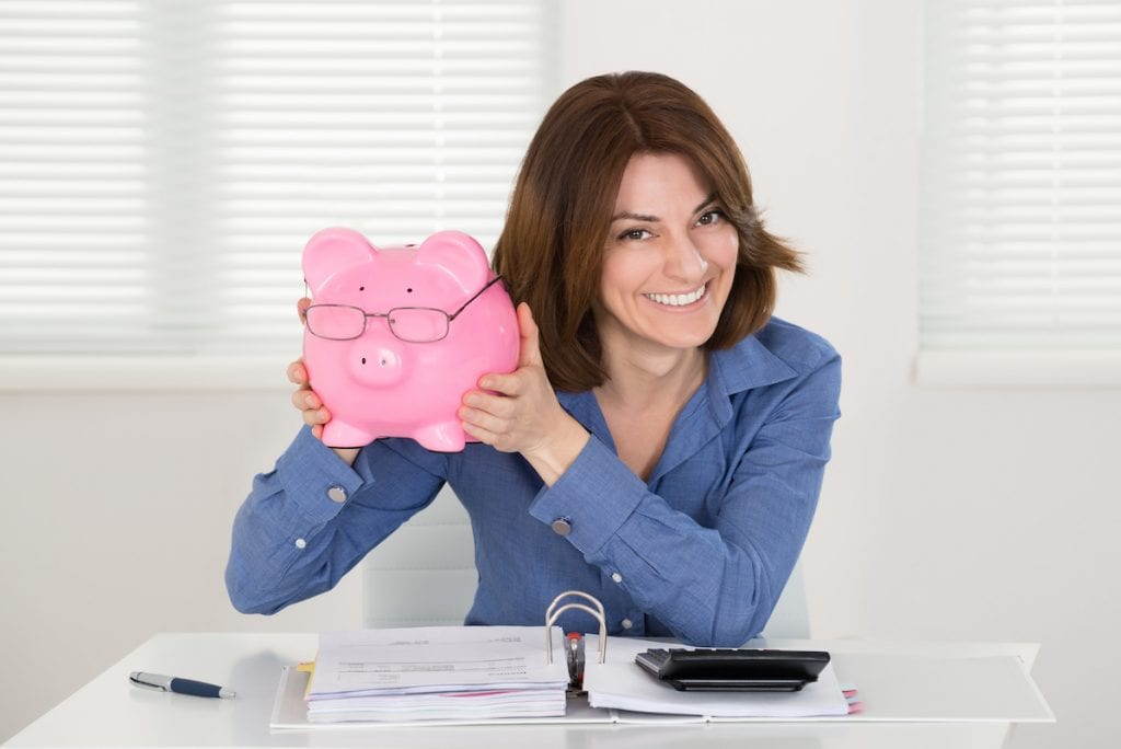 Happy Young Businesswoman Holding Piggybank With Document On Desk At Home