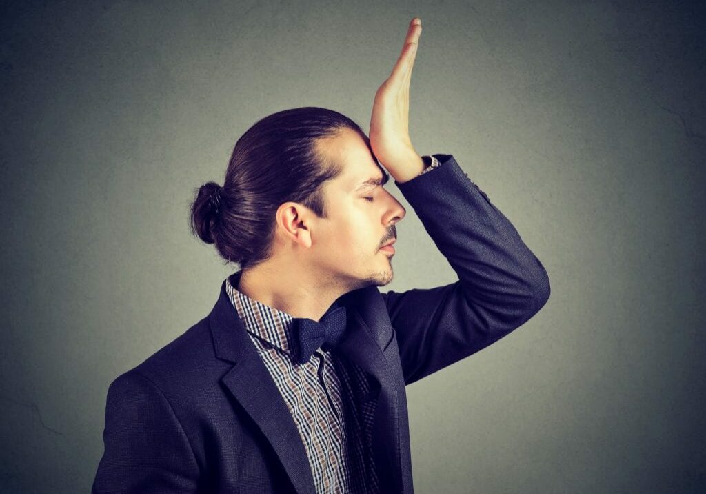 Young elegant man slapping forehead upset with foolish mistake while posing on gray.